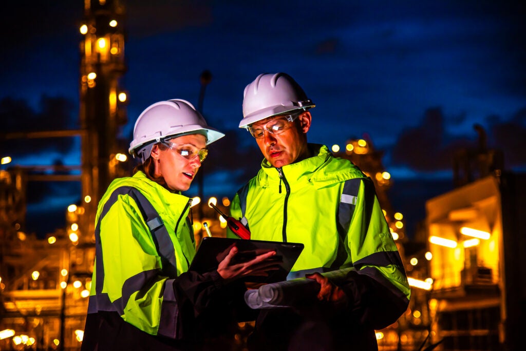 Octave_Hexagon Team of construction engineers in uniforms and helmets examine project with tablet Industrial plant or oil refinery background at night.